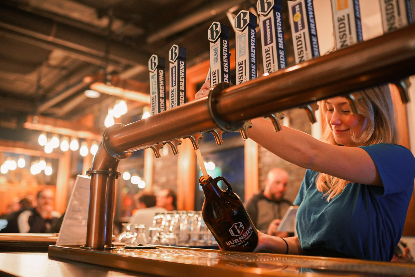A woman in a blue shirt pours beer from a copper tap system into a dark brown growler at a bar. Multiple taps with visible 'Burnside Brewing' branding are mounted above the counter, and the background shows a softly blurred pub interior with lights and patrons.