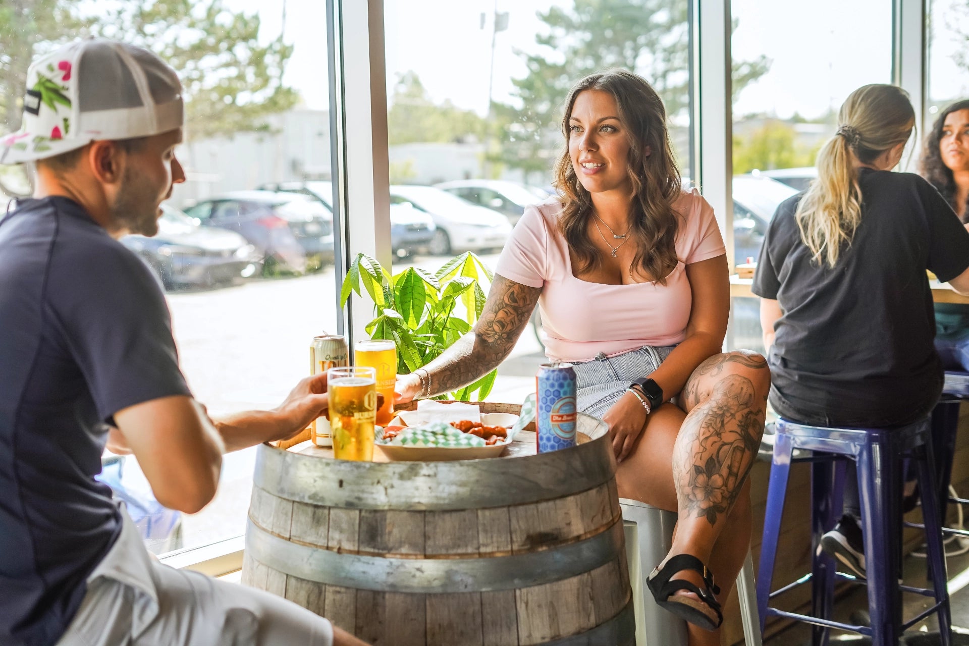 A man and woman sitting around a barrel table in Burnside Brewing with drinks and food.The woman has a sleeve of tattoos down one arm and leg.